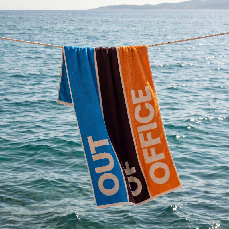 Dzintars Out Of Office premium cotton beach towel 90x170cm hanging on a rope over the sea – colour-block design in blue, burgundy and orange