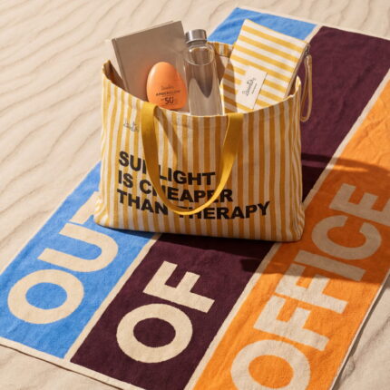 Dzintars Out Of Office beach towel 90x170cm laid on sand with striped tote bag and Amberglow SPF 50+ face moisturizer – premium cotton summer lifestyle